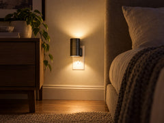 A black Aoralight at night plugged into a wall outlet, shown at its real size beside the outlet plate, giving off a soft